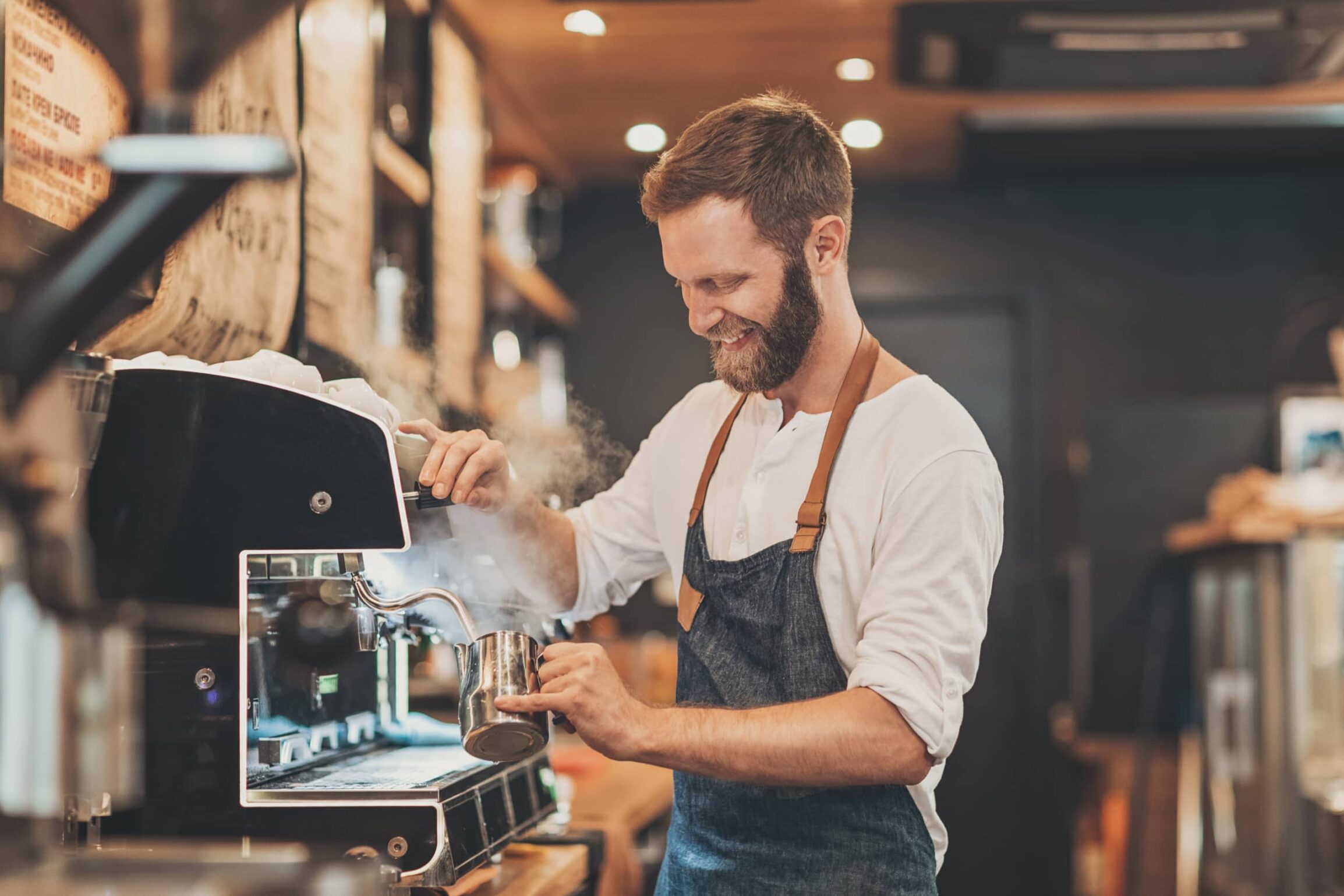 Smiling barista in an apron steams milk with an espresso machine at a cozy café. Warm lighting and wood accents create an inviting atmosphere.