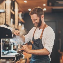 Smiling barista in an apron steams milk with an espresso machine at a cozy café. Warm lighting and wood accents create an inviting atmosphere.