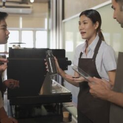Four baristas in a cafe, gathered around an espresso machine. One is holding a jar of coffee beans, explaining, others listen attentively. Energetic atmosphere.