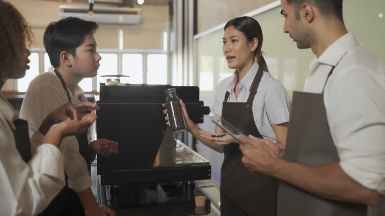 Four baristas in a cafe, gathered around an espresso machine. One is holding a jar of coffee beans, explaining, others listen attentively. Energetic atmosphere.