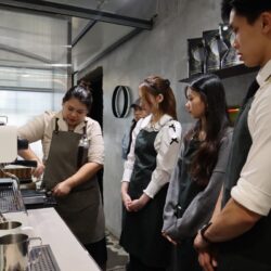 Barista in apron, demonstrating espresso machine use to attentive group of young adults, evoking a learning atmosphere in a modern café setting.