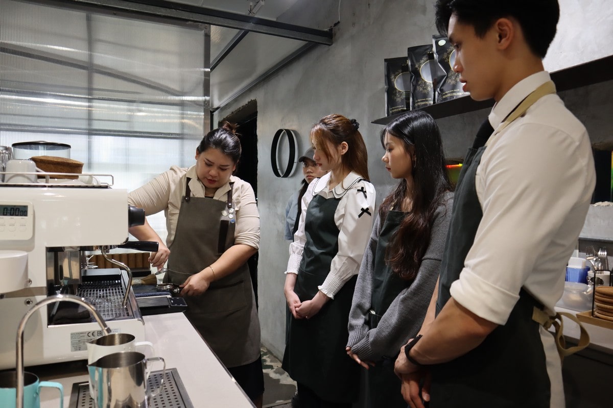 Barista in apron, demonstrating espresso machine use to attentive group of young adults, evoking a learning atmosphere in a modern café setting.