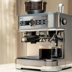 A sleek silver espresso machine with a built-in grinder sits on a kitchen counter. It brews fresh coffee into a clear glass cup. Sunlight filters through a window, adding warmth.