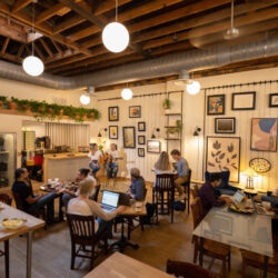 Cozy cafe with wooden decor, hanging plants, and framed art. People work on laptops and chat, creating a warm, collaborative atmosphere.
