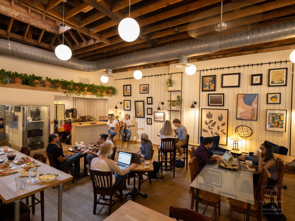 Cozy cafe with wooden decor, hanging plants, and framed art. People work on laptops and chat, creating a warm, collaborative atmosphere.