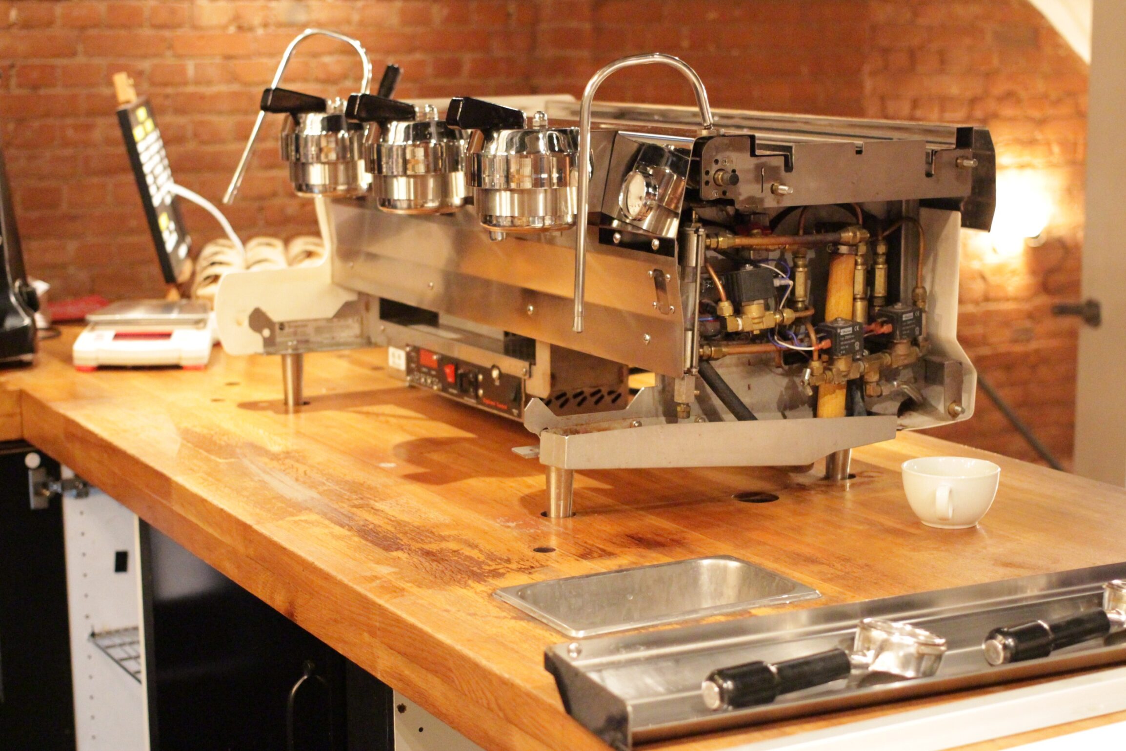 Commercial espresso machine on a wooden cafe counter, partially disassembled showing internal components. Warm lighting with exposed brick wall creates a cozy ambiance.
