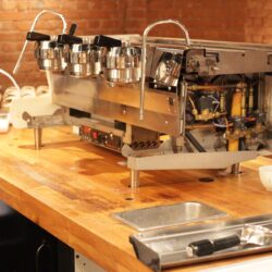 Commercial espresso machine on a wooden cafe counter, partially disassembled showing internal components. Warm lighting with exposed brick wall creates a cozy ambiance.