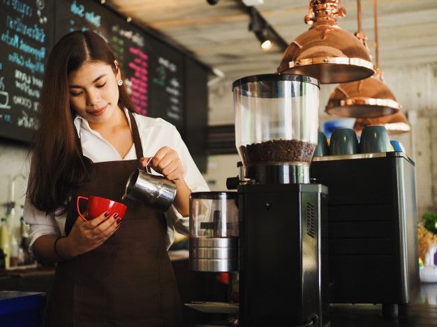 Barista in a brown apron pours milk into a red cup at a coffee shop. She stands by a coffee machine with a warm, focused expression. Copper lights hang above.